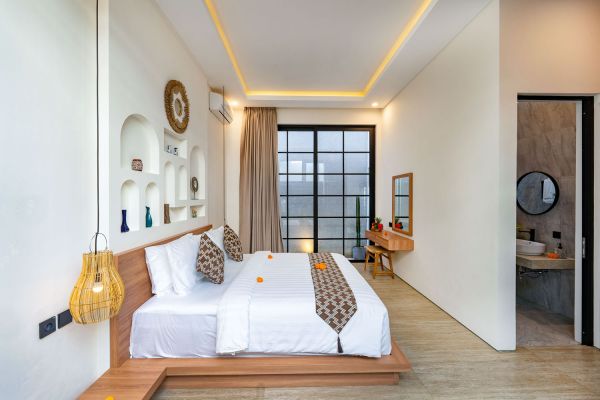 Spacious Balinese bedroom with wooden platform bed, white linens, orange accents, woven pendant light, and large curtained window.